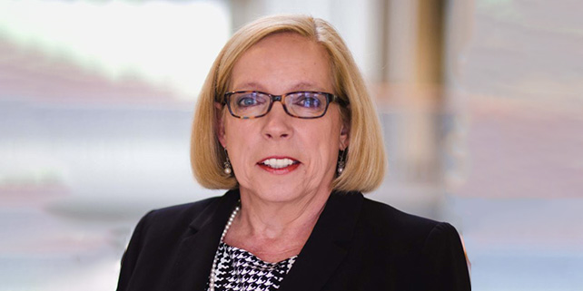 Mary Holcomb - woman with blond hair, wearing glasses, a dark suit jacket over a herringbone topand pearls